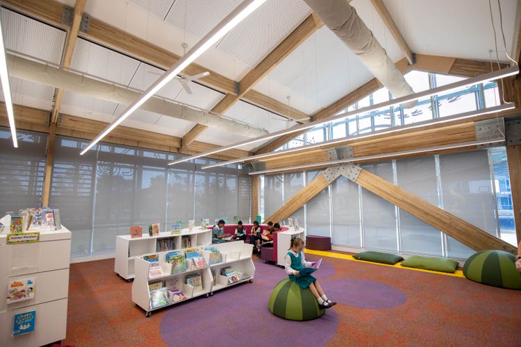 A view of the library space. Tall ceilings with exposed wooden beams criss-cross the structure. Shelving and seating spaces are scattered around.