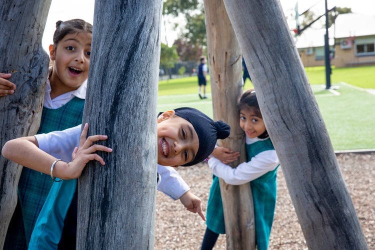 Two girls and a boy are peeking out from between the beams of a wooden play structure.