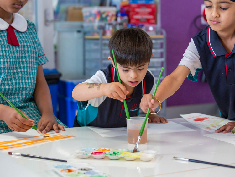 Three students painting. The student in the centre is in full focus dipping his paintbrush in the water. The student to the right is partially out of frame and dipping his paintbrush into a tray. The student to the left is almost fully out of frame and is painting the sheet of paper yellow.