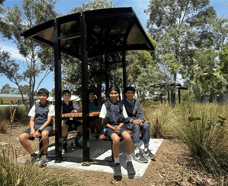 Five students sitting on a shaded picnic bench within a landscaped bush garden