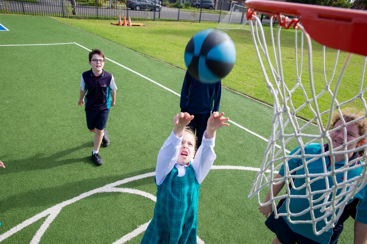 Three students playing basketball. A girl throws a blue and black ball upwards, the net in the foreground