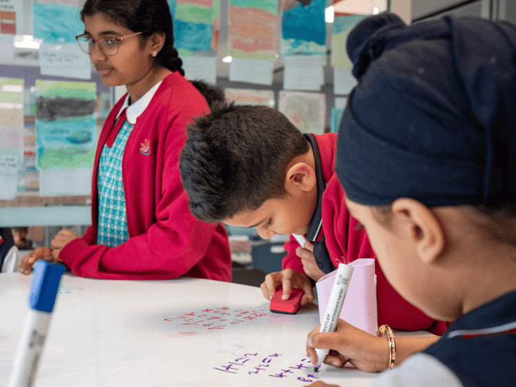 Three students at a whiteboard table writing mathematical equations. One girl and two boys.