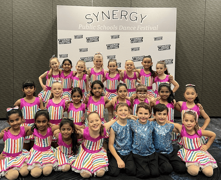 A junior dance team posing for a photo in front of the Synergy sign