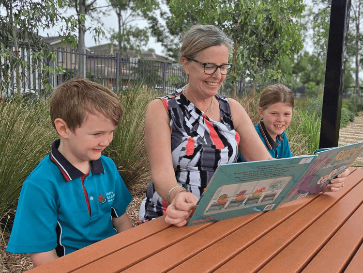The principal, Mrs Lisa Crawford, reading to two stuents, a boy and a girl. They are sitting either side of the principal at a picnic table outdoors.