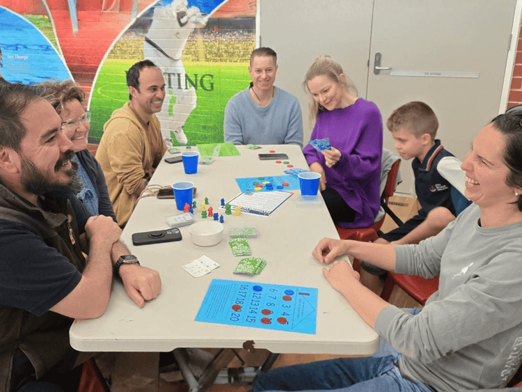 6 parents are seated around a table with a student demonstrating some maths games. There are resources on the table.